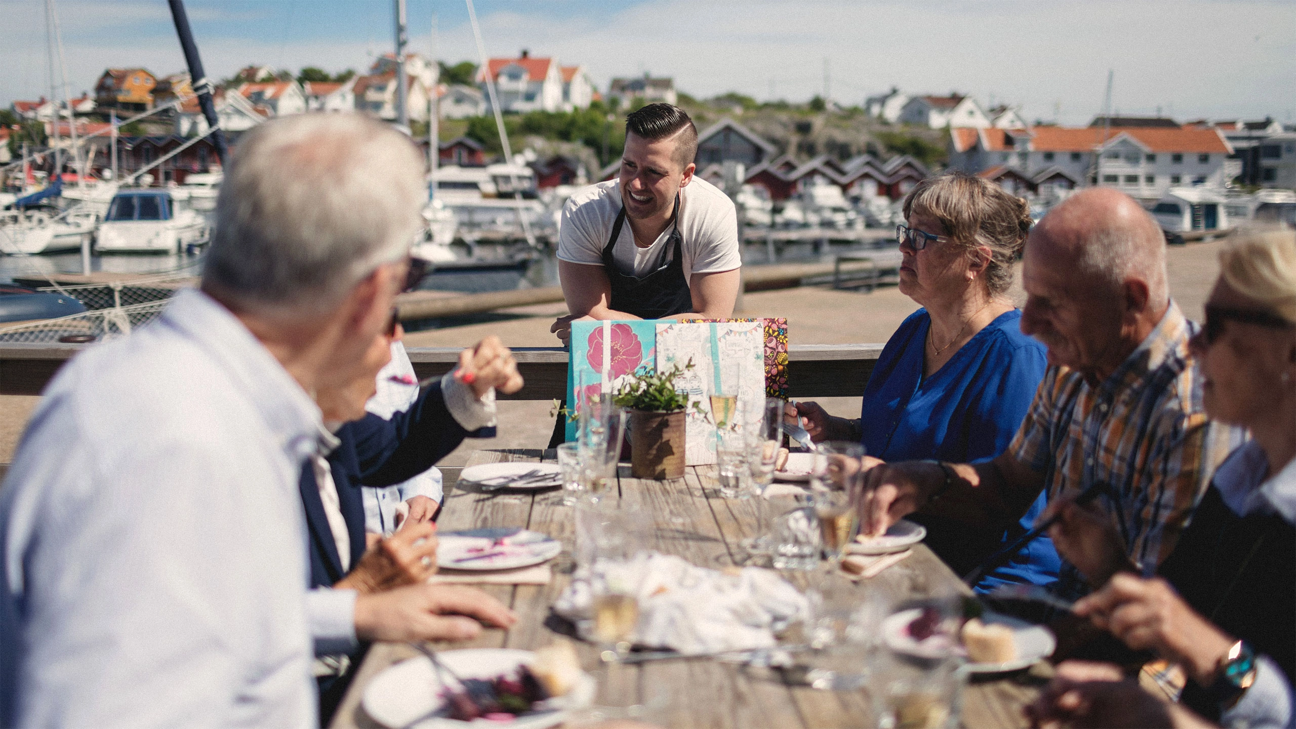 Tjener taler med mennesker, der sidder og spiser ved et bord udenfor med havn og huse i baggrunden